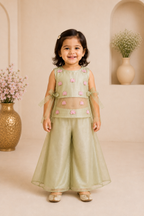 Young girl in a green traditional outfit standing in a decorated room with vases and flowers.