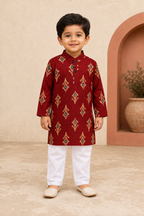 Young boy wearing a maroon kurta with white pants standing against a beige wall.