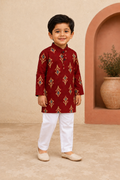 Young boy wearing a maroon kurta with white pants standing side ways against a beige wall.