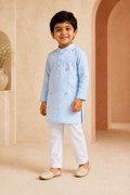 Young boy in a light blue kurta with white pants standing in a beige room.