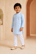 Young boy in a light blue kurta with white pants standing in a beige room.